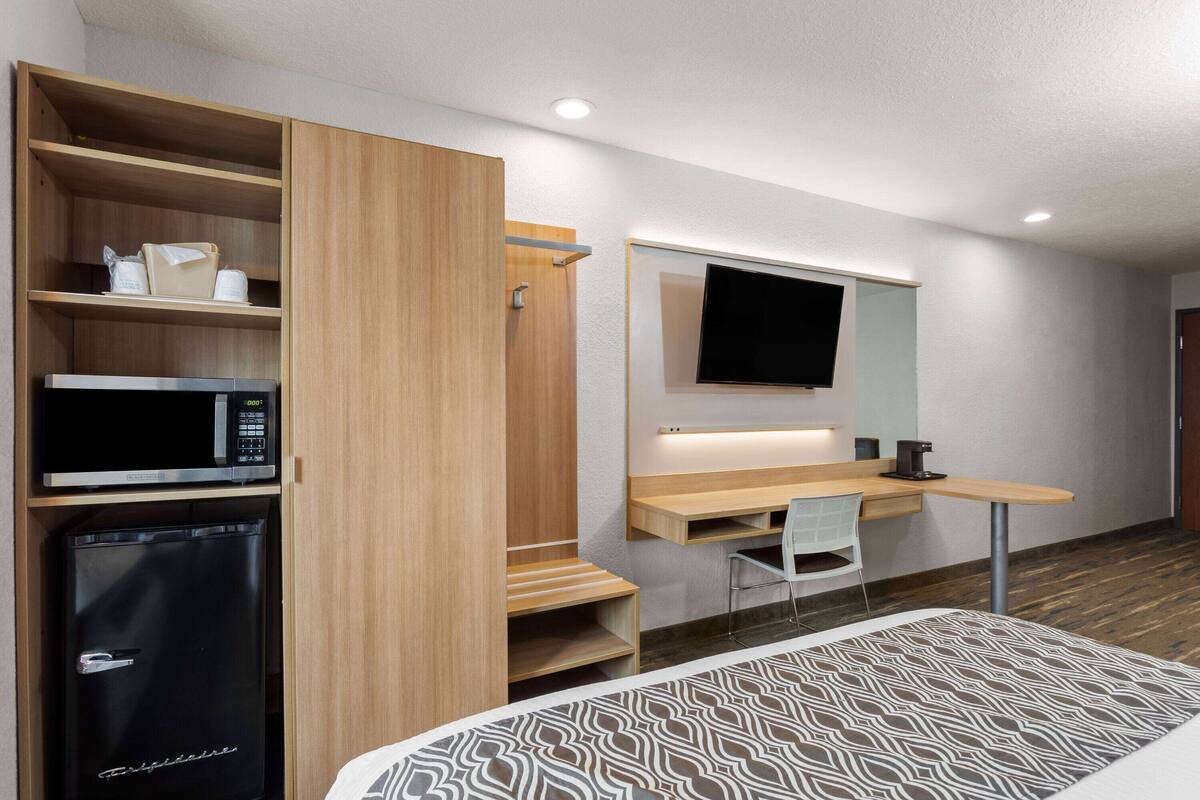 Habitación de hotel pequeña con cama, escritorio con silla, microondas, refrigerador y televisor montado en la pared.