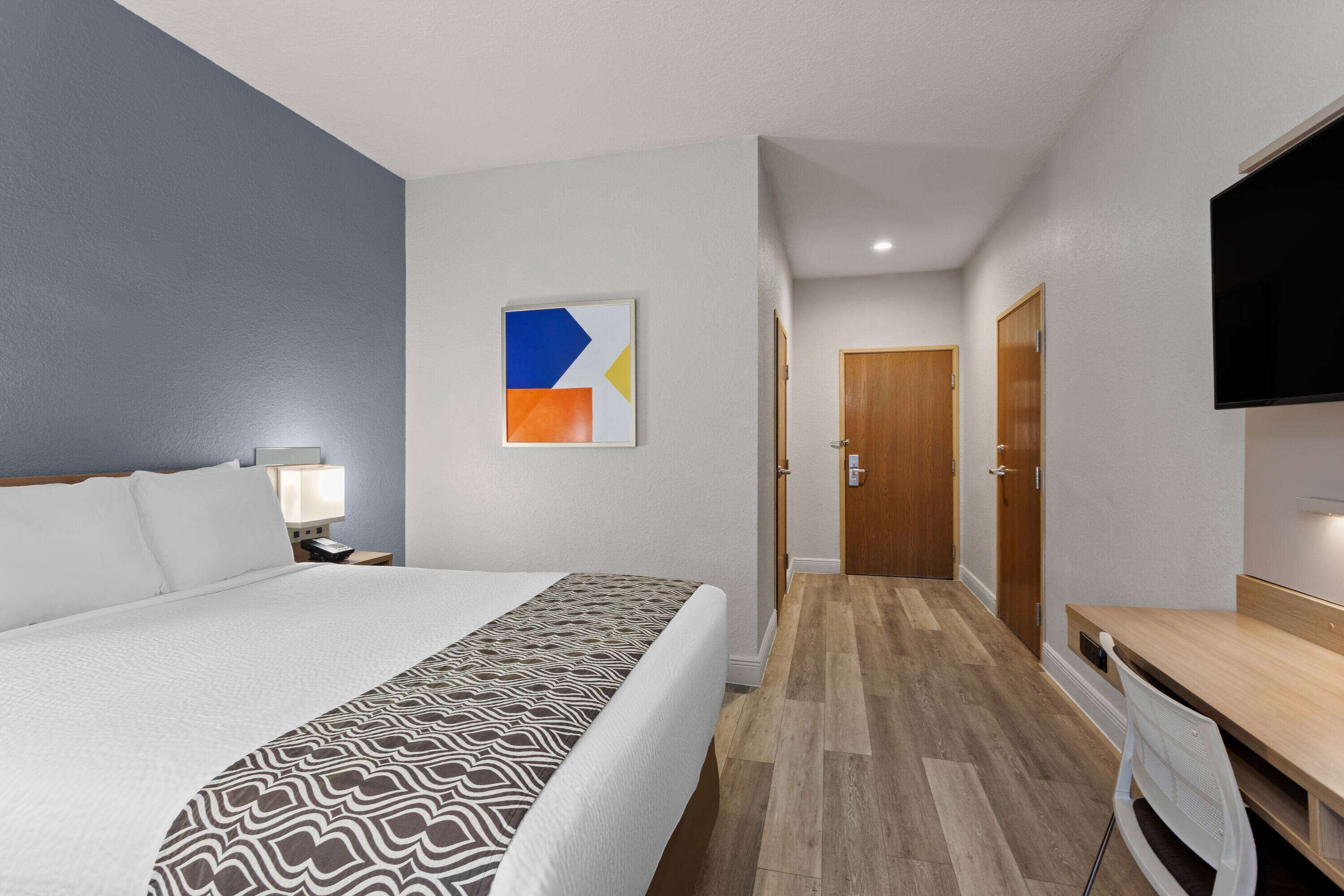 A hotel room with a large bed, a desk, a chair, a TV, a bedside table with a lamp, and a door leading to another room.