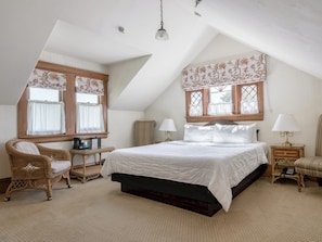 Un dormitorio con una cama, dos mesitas de noche, una silla, una ventana con cortinas y una lámpara colgante.