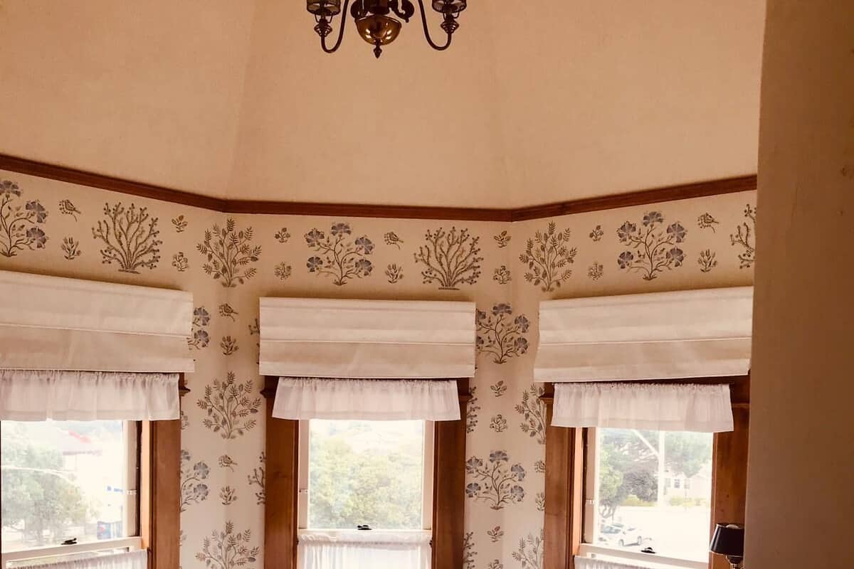 A bedroom with a large bed, two windows with floral curtains, a chandelier, and a view of the outside.