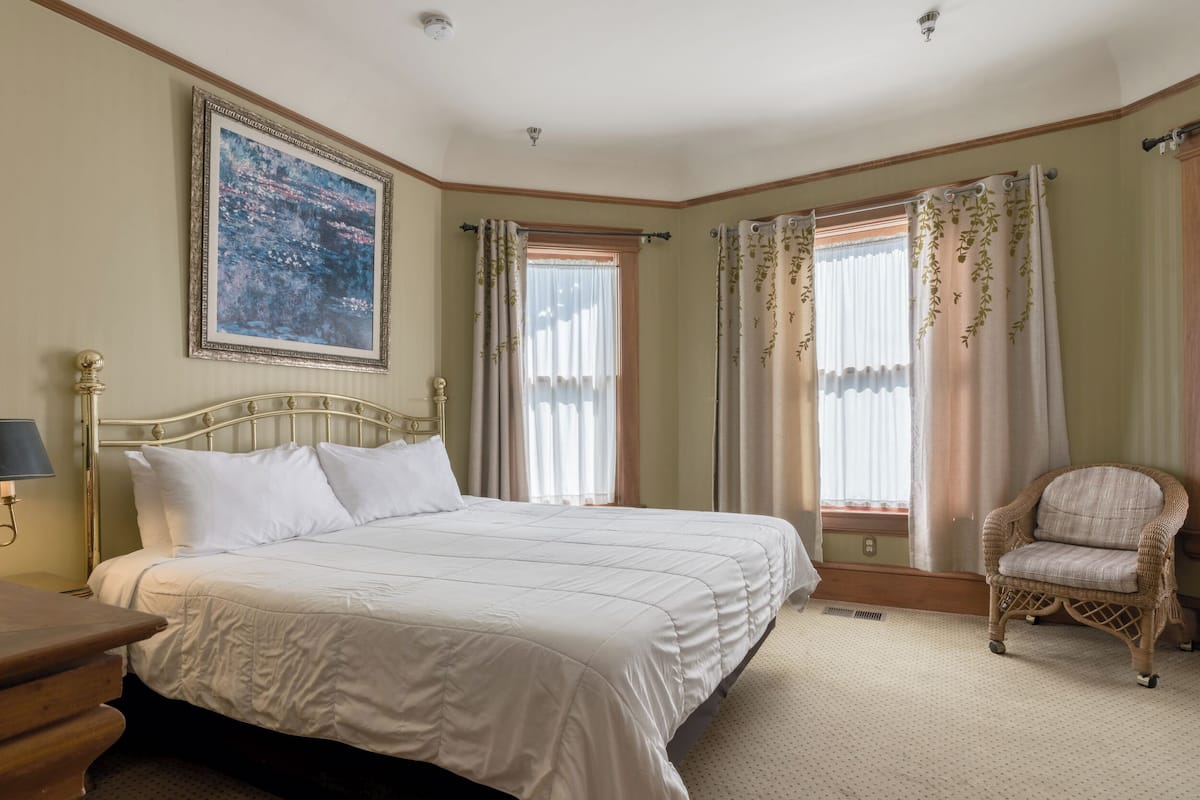 A bedroom with a bed, a chair, a painting, and windows with curtains.