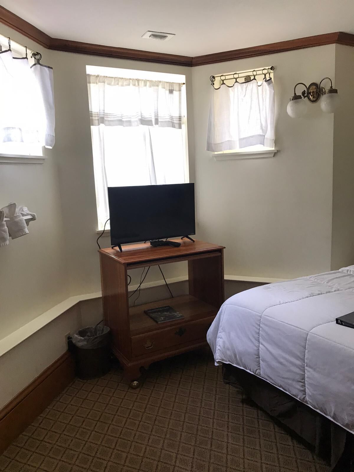 A hotel room with a bed, a wooden TV stand with a remote control, a small trash bin, and a window with curtains.