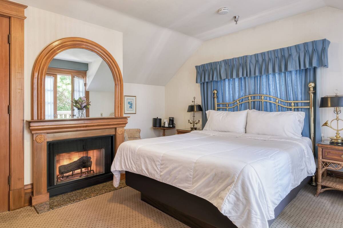 A bedroom with a fireplace, a large bed, a nightstand, a lamp, and a window with curtains.