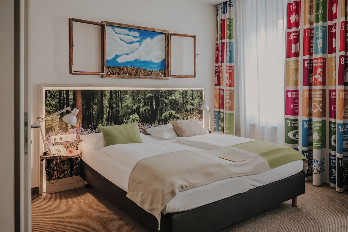 A neatly made bed with a headboard featuring a forest print, a bedside table with a lamp, and a wall with a framed picture and a curtain with a UN theme.