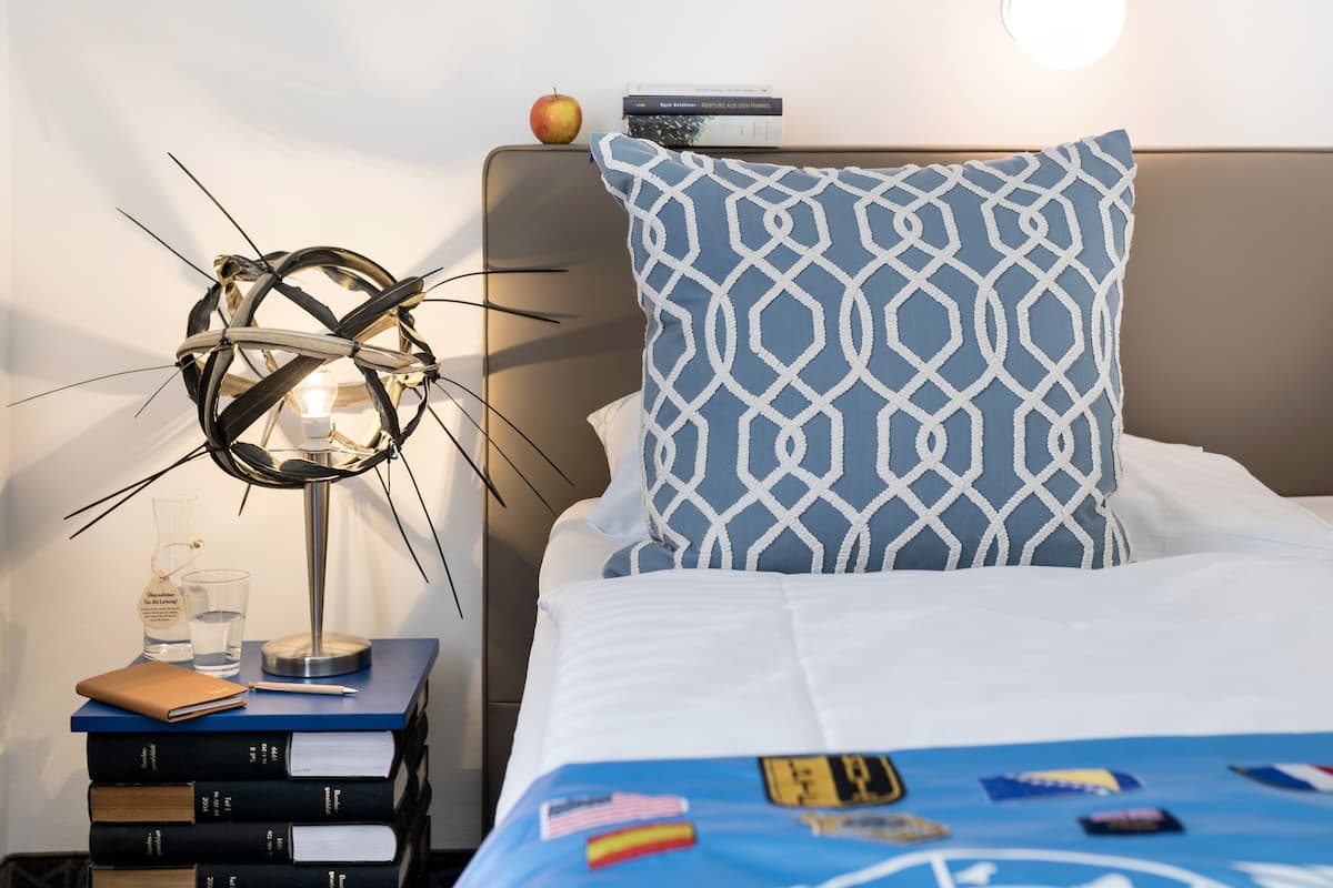 A neatly made bed with a patterned pillow, a bedside table with books and a lamp, and a small apple on the headboard.