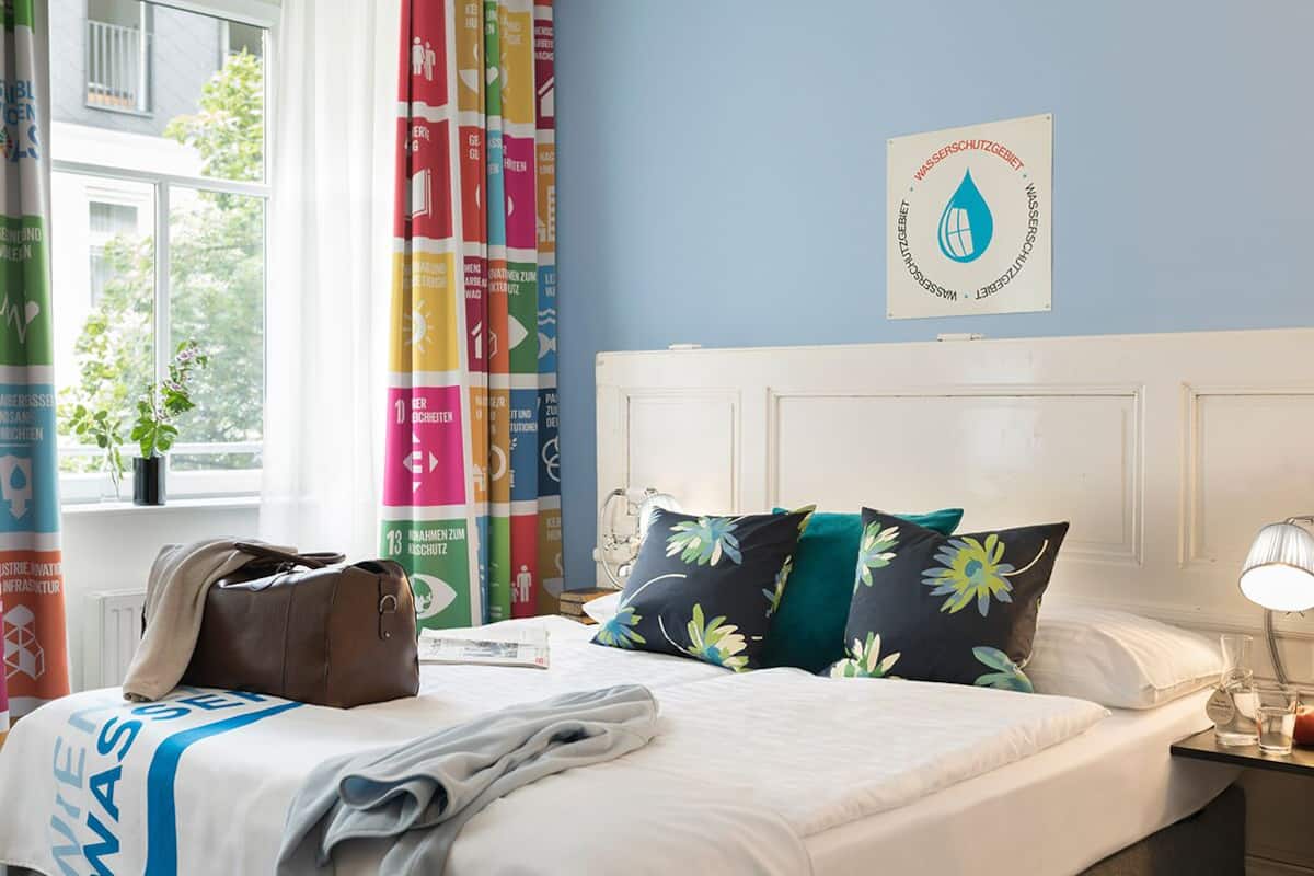 A hotel room with a bed, pillows, a nightstand, a lamp, a window with curtains, and a wall sign.