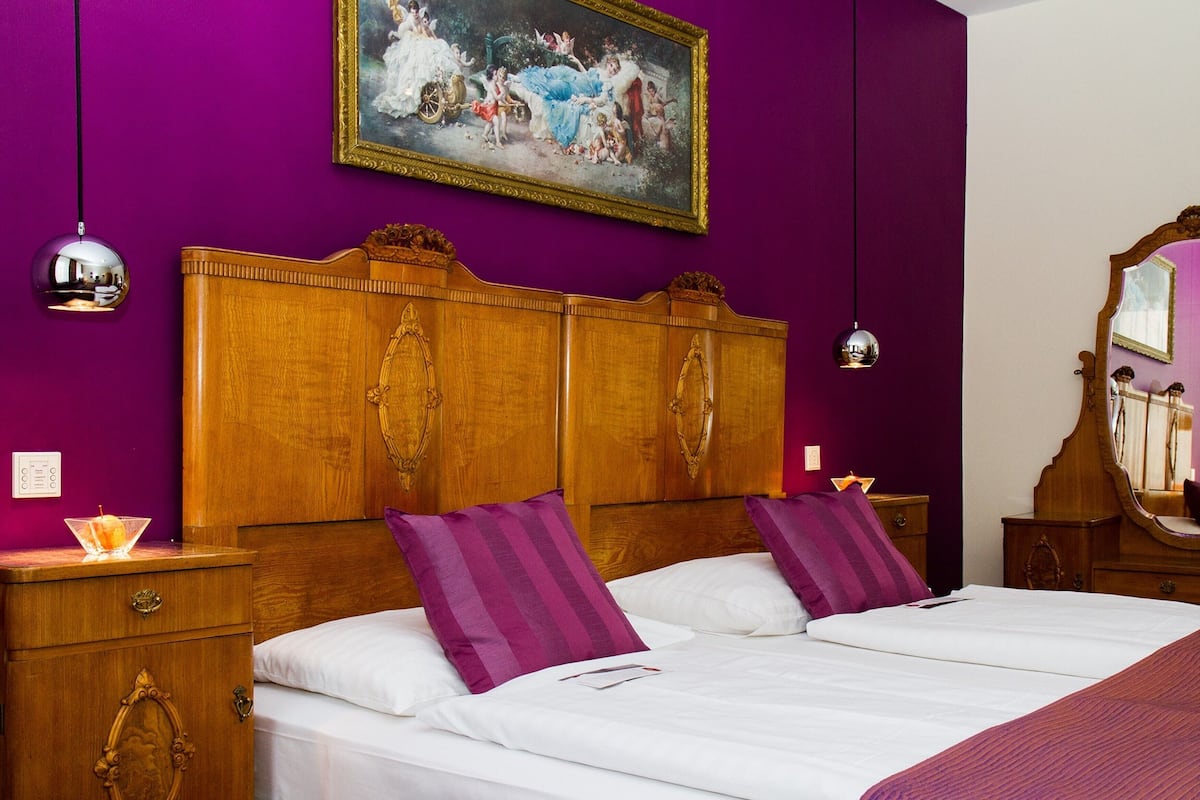 A room with a wooden bed, purple walls, a classic painting, and a wooden dresser.