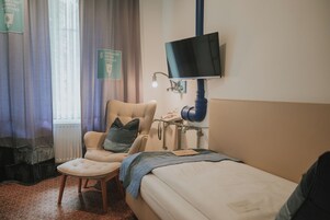 Een hotelkamer met een bed, een stoel, een bureau met een lamp, een aan de muur bevestigde televisie en een raam met gordijnen.