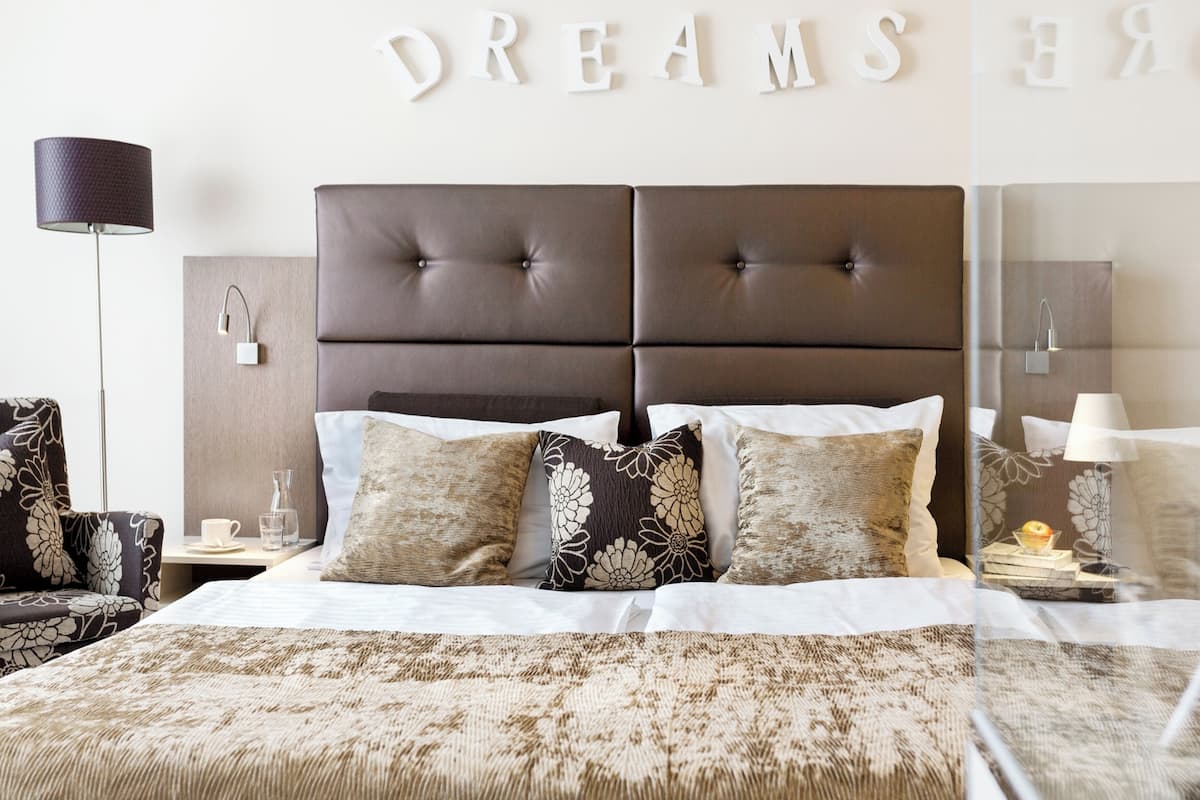 A bedroom with a tufted headboard, a bed with patterned bedding, a side table with a lamp, and a wall-mounted mirror.