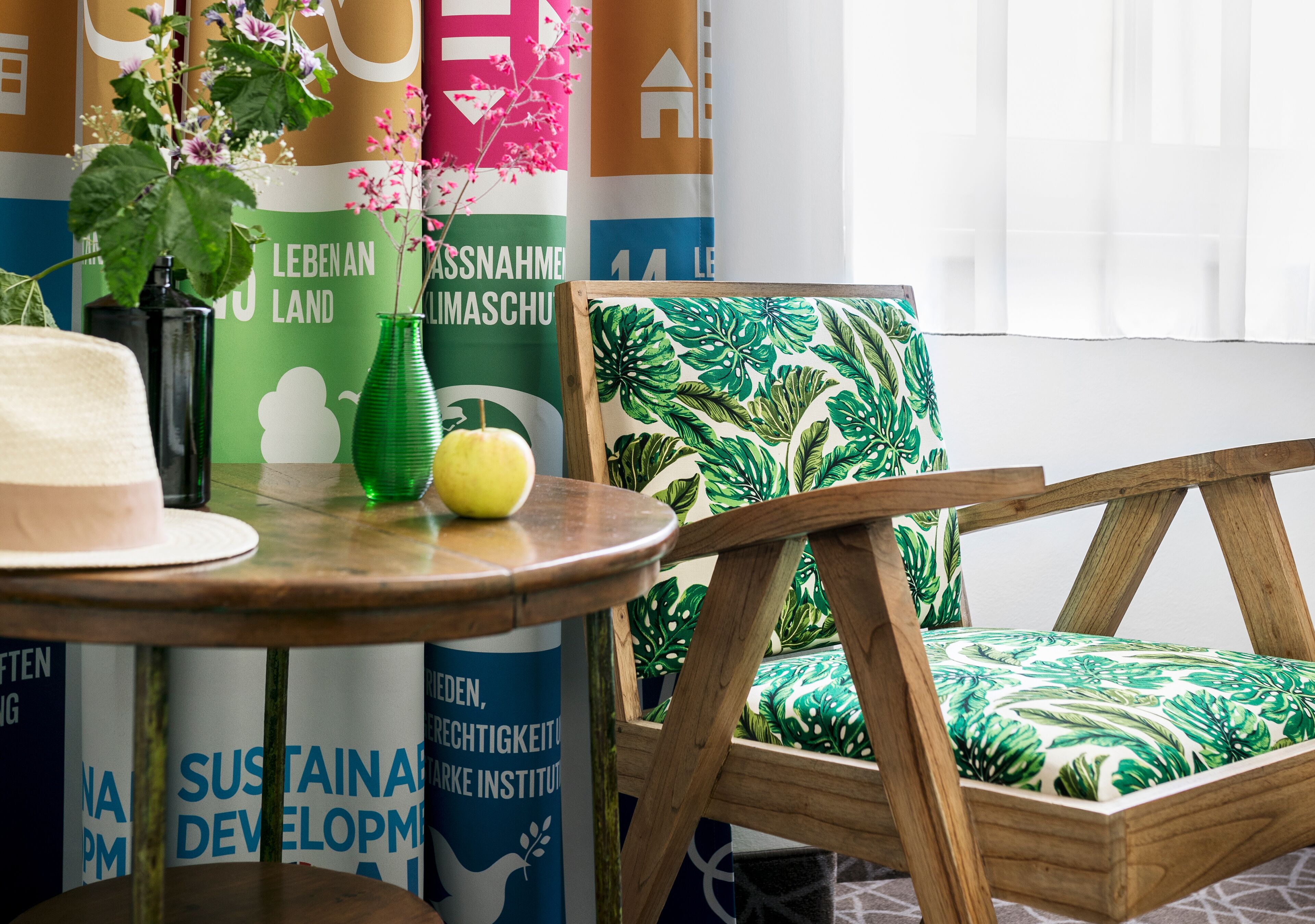 Una habitación con una mesa redonda de madera, un jarrón verde y una manzana amarilla. Hay carteles en la pared y una silla verde con estampado.