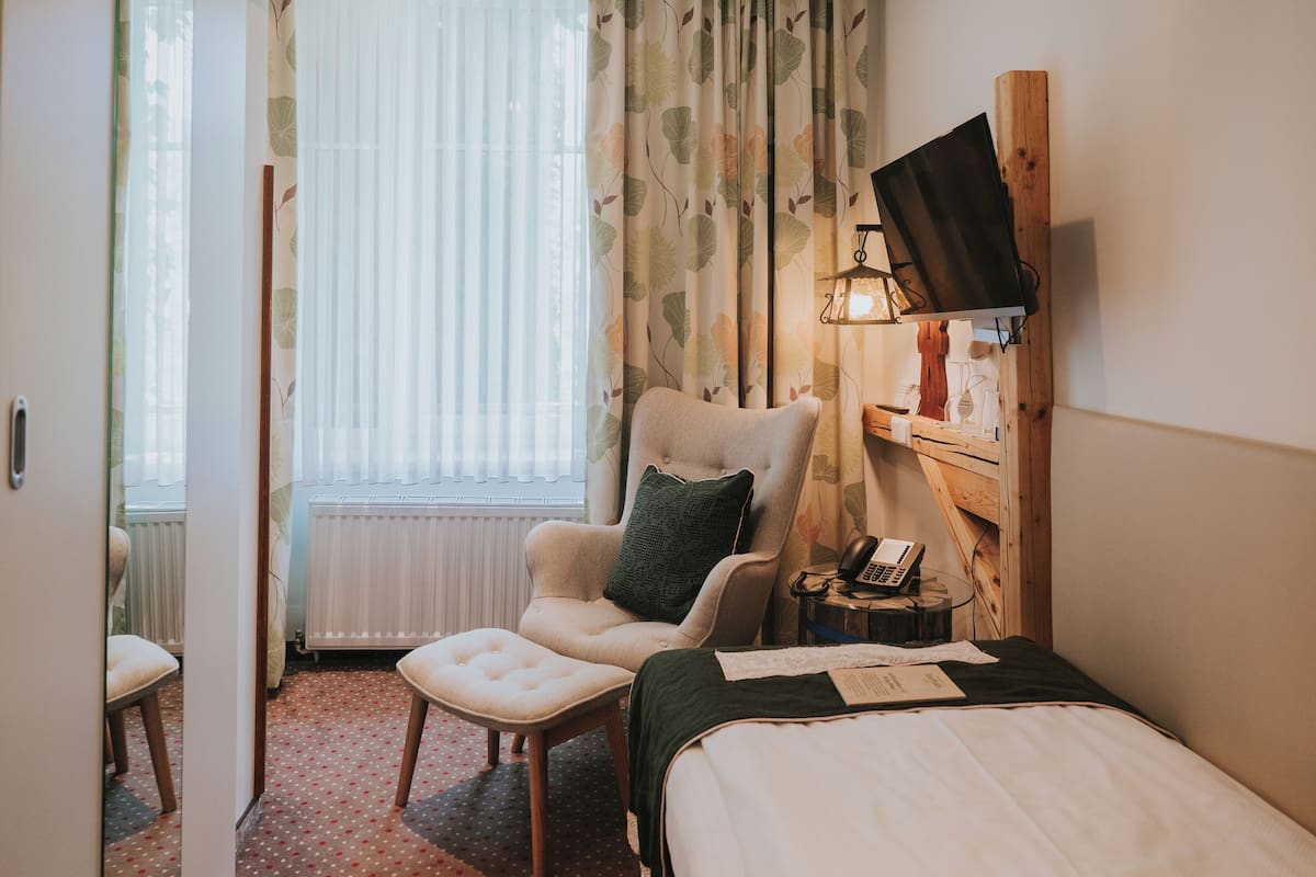 A hotel room with a bed, a chair, a desk with a telephone, and a window with curtains.