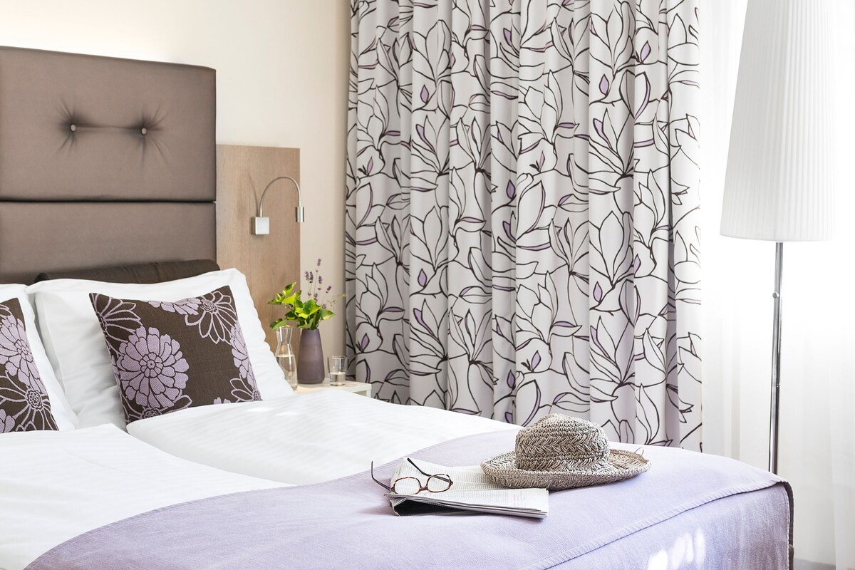 A neatly made bed with a headboard, a bedside table with a vase and a lamp, and a window with curtains.