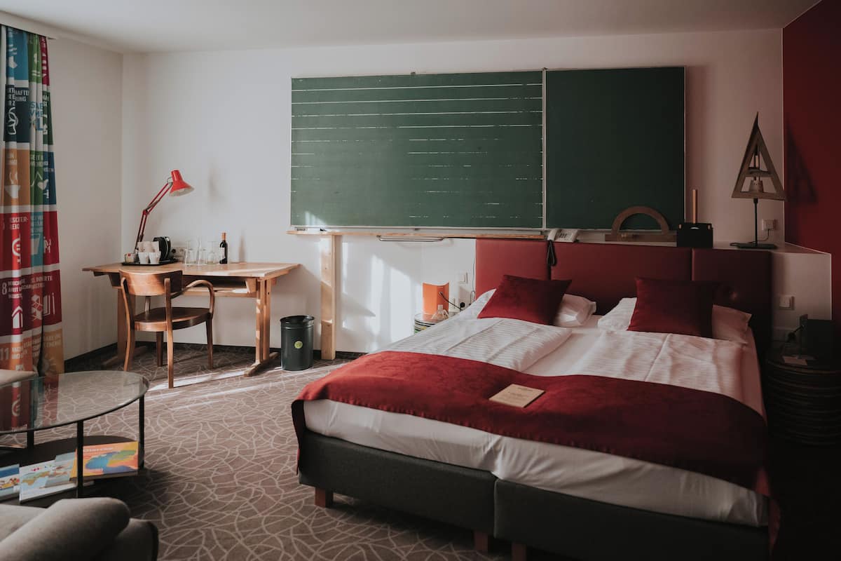 A hotel room with a bed, a desk, a chair, a bookshelf, and a chalkboard.