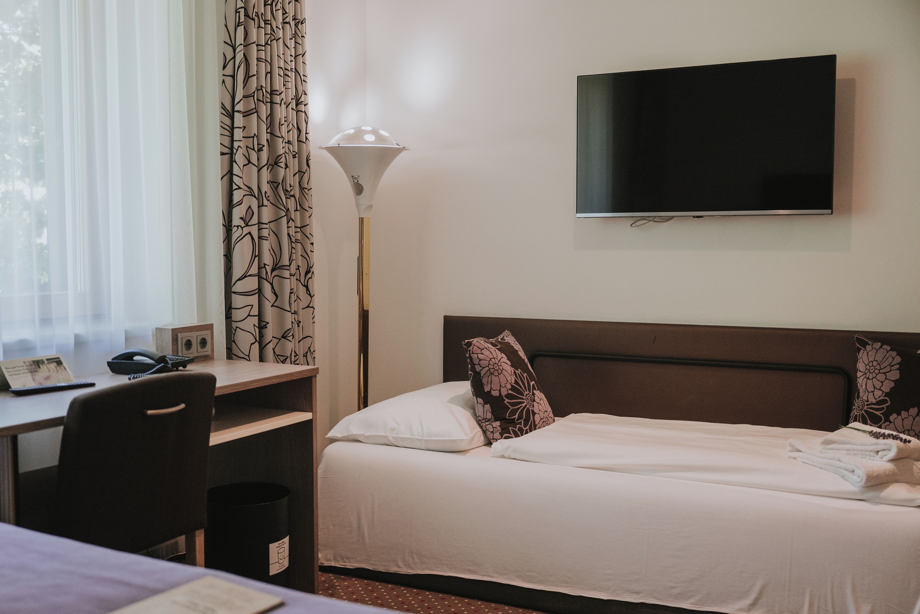 A hotel room with a bed, a desk with a chair, a lamp, and a television on the wall.