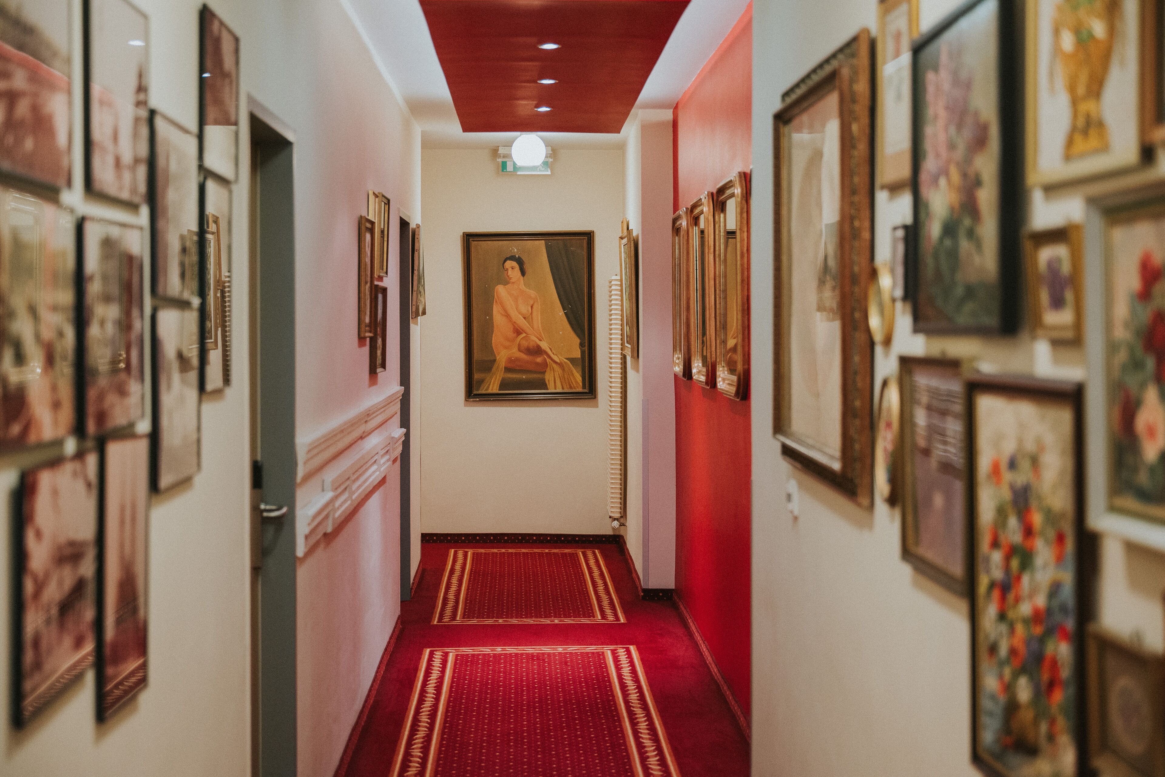 A hallway with red carpet, framed artwork on the walls, and a central painting.