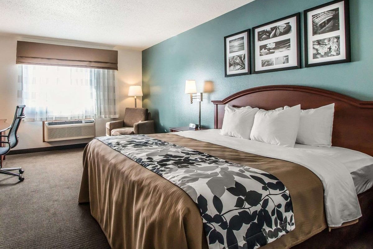 A hotel room with a bed, a desk, a chair, a window with curtains, and wall-mounted lamps.