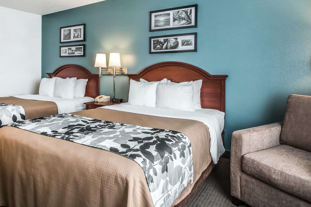 A hotel room with two beds, a chair, and wall-mounted lamps.
