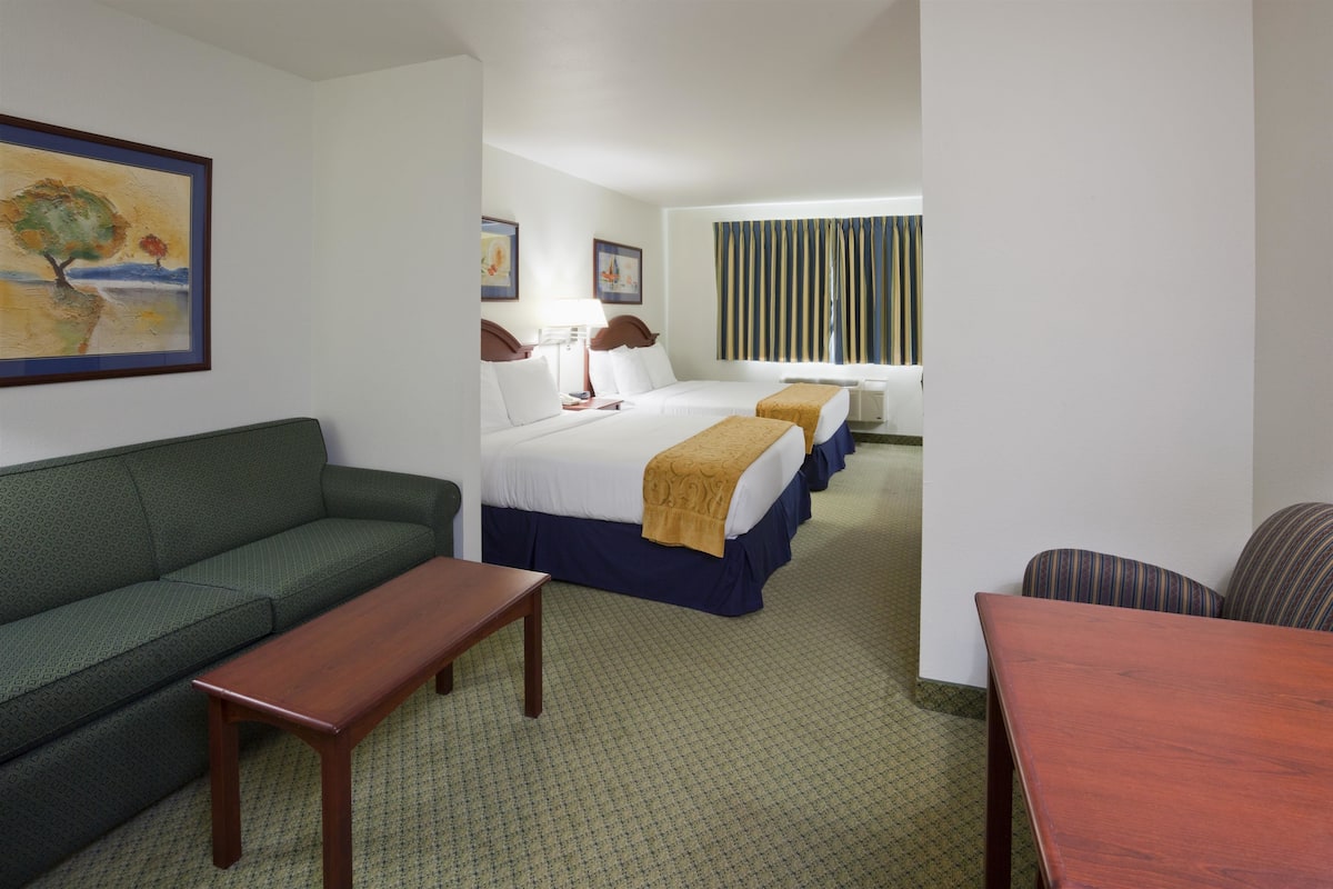 A hotel room with a green sofa, a wooden table, two beds with blue and white bedding, and a window with striped curtains.