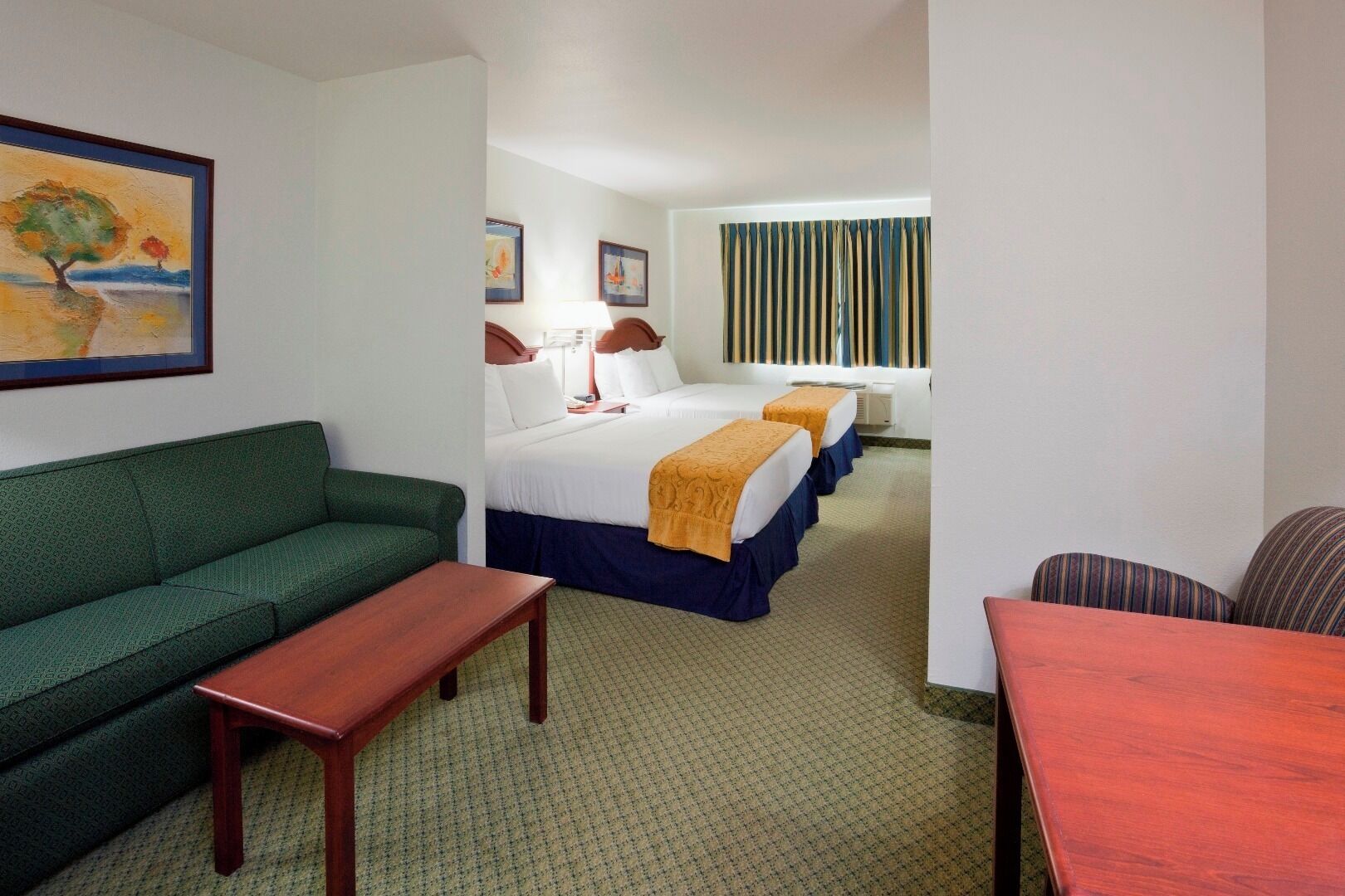 A hotel room with a green sofa, a wooden table, two beds with blue and orange bedspreads, and a painting on the wall.