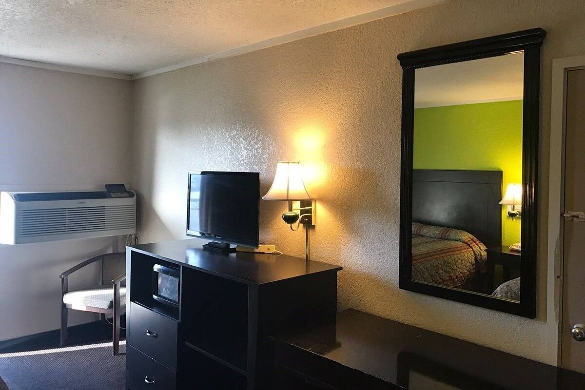 A hotel room with a desk, television, lamp, and a large mirror reflecting a bed.