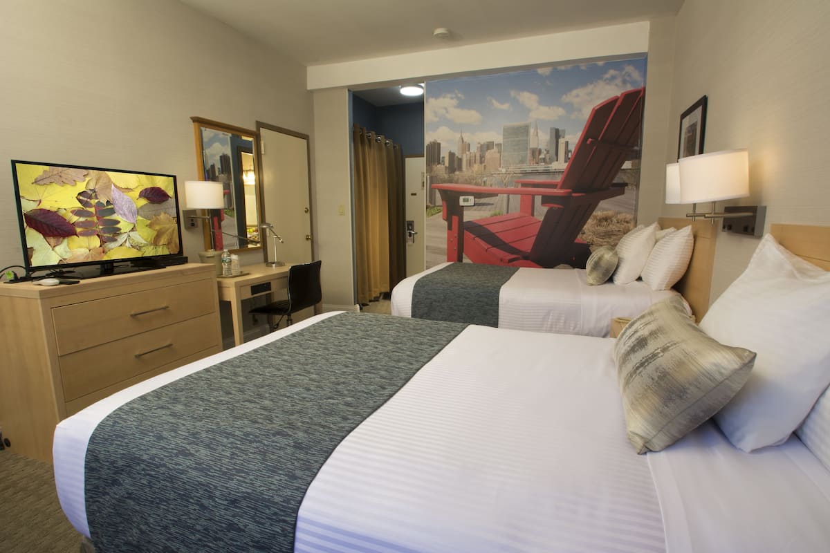 Une chambre d’hôtel avec deux lits, un bureau, une chaise, un téléviseur et une fresque représentant un paysage urbain sur le mur.