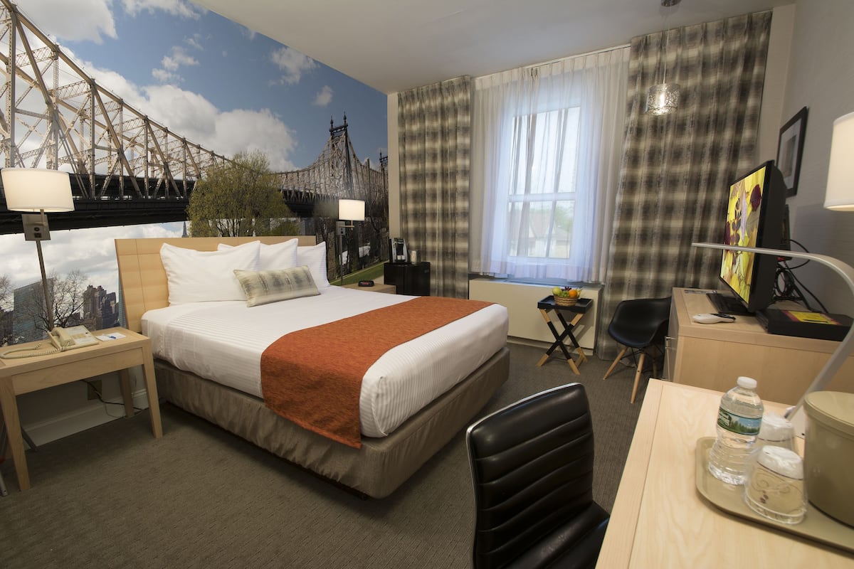 Une chambre d’hôtel avec un lit, un bureau, une chaise, une lampe de bureau, une bouteille d’eau et un tableau au mur.