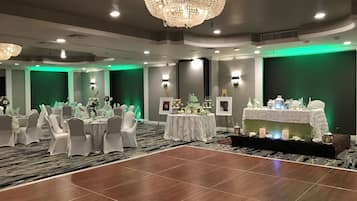 Une salle de banquet avec des tables rondes, des chaises, une piste de danse et une scène où sont disposés un gâteau et des photos.