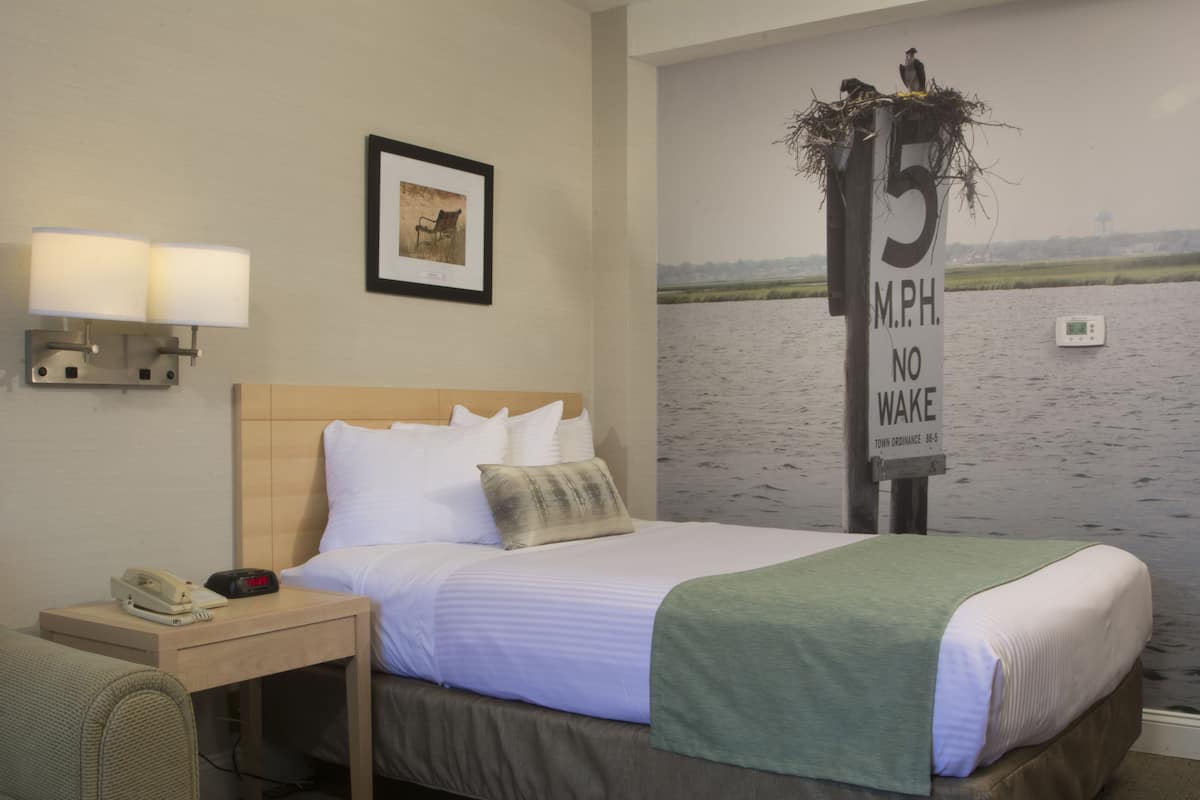 Une chambre d’hôtel comprenant un lit, une table de chevet avec un téléphone et un réveil, une lampe fixée au mur, un tableau encadré et une fresque représentant une rivière avec un panneau indiquant une vitesse maximale de 15 miles par heure.