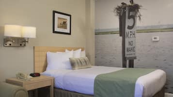 A hotel room with a bed, a nightstand with a phone and alarm clock, a wall-mounted lamp, a framed picture, and a mural of a river with a sign indicating a speed limit of 15 mph.