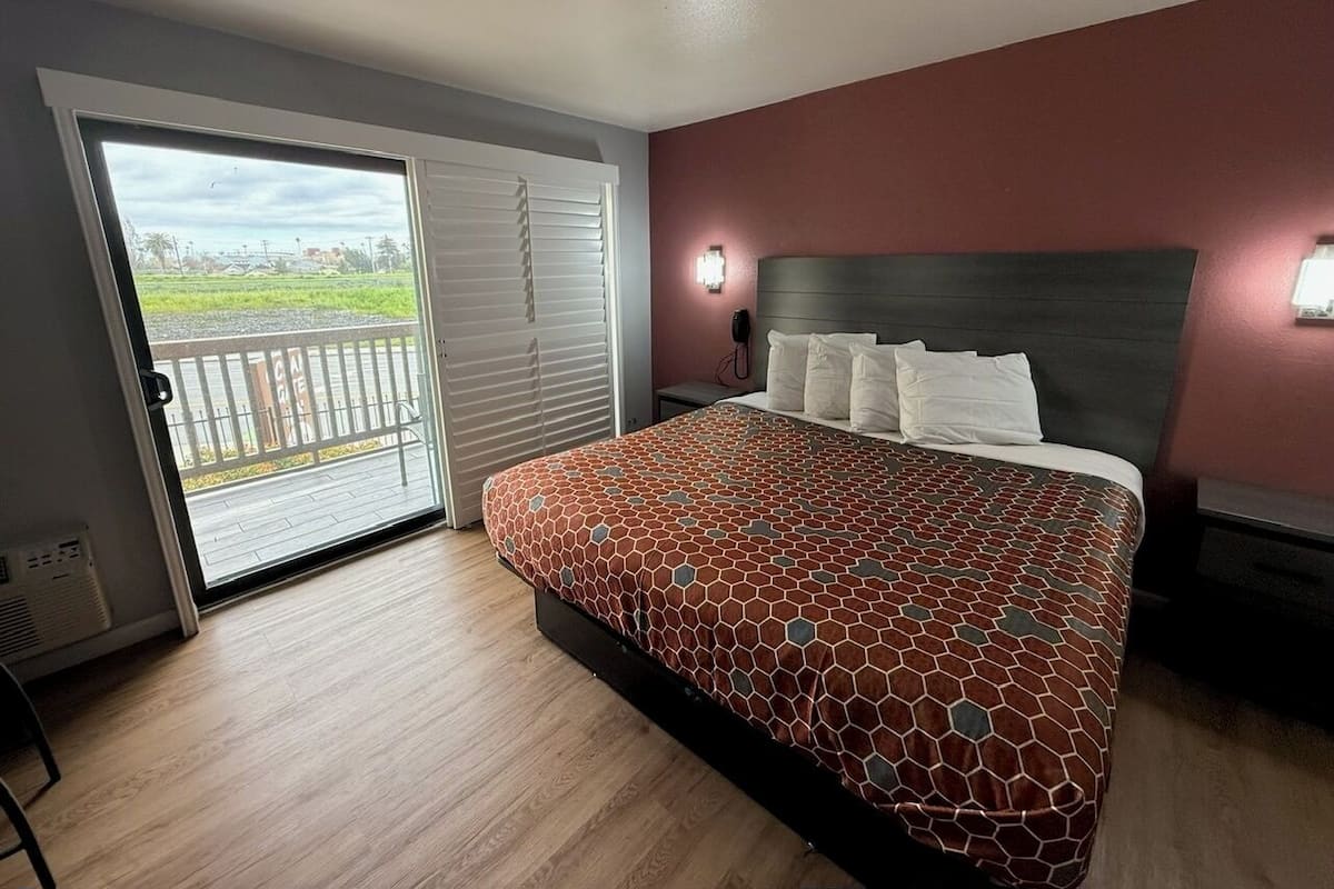 A bedroom with a large bed, a sliding glass door to a balcony, and a nightstand with a lamp.