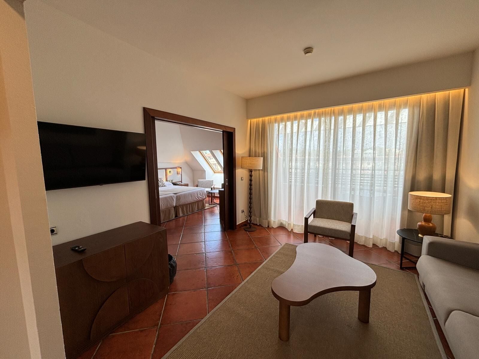 A hotel room with a bed, a TV, a desk, a chair, and a large window with curtains.