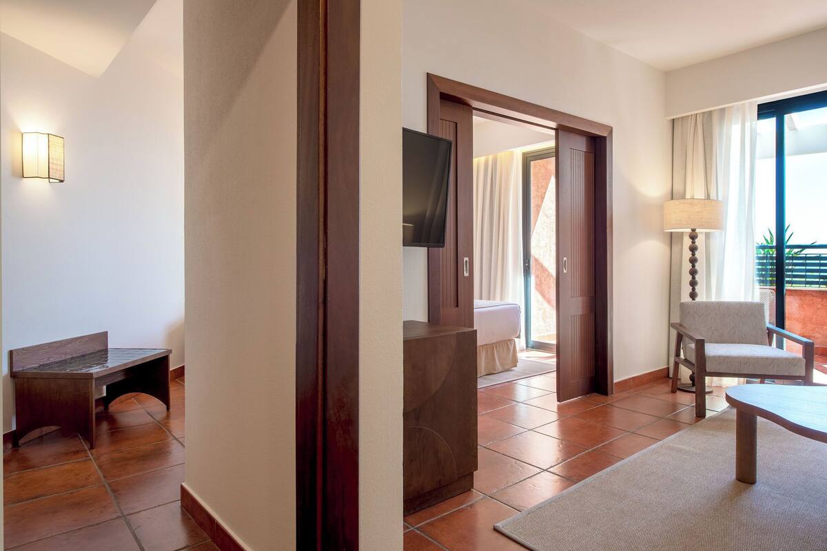Een hotelkamer met een houten bankje, een flatscreen televisie aan de muur, een stoel en een groot raam met gordijnen.