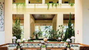 A spacious lobby with a central seating area, surrounded by lush greenery and a high ceiling with a patterned design.