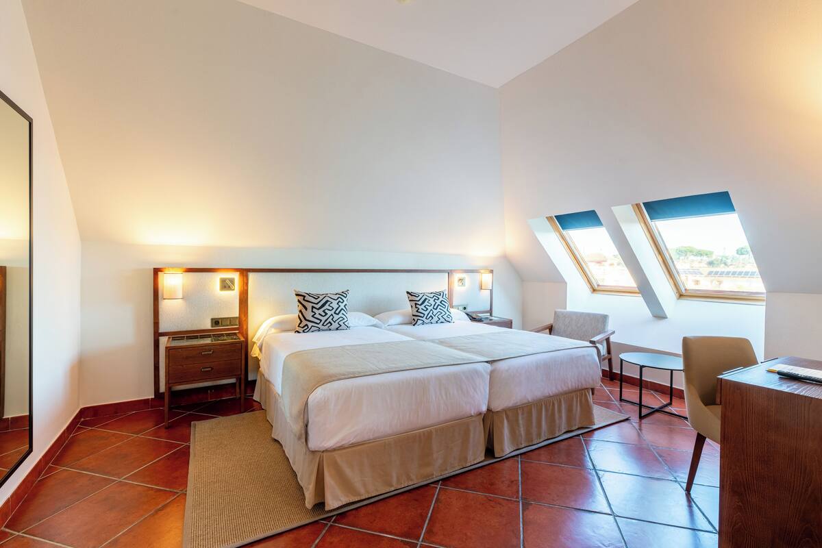 Een slaapkamer met een groot bed, twee nachtkastjes, een bureau en een stoel. Er zijn twee ramen die zorgen voor natuurlijk licht.