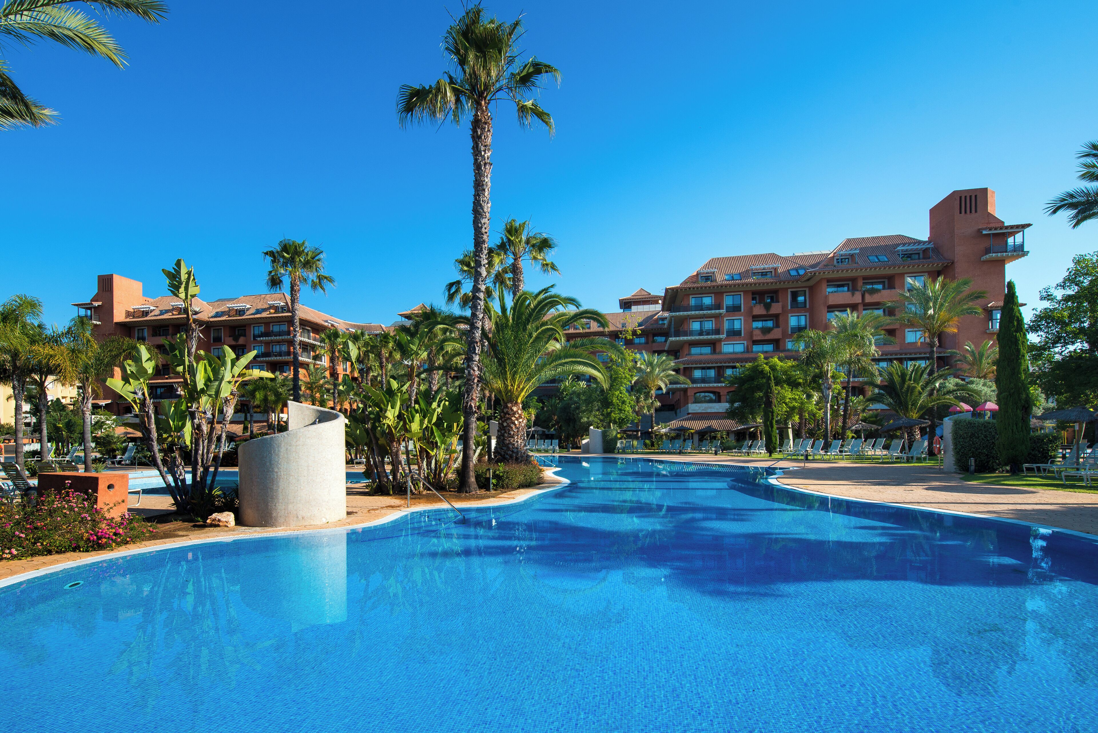A large swimming pool surrounded by palm trees and a hotel building with multiple balconies.