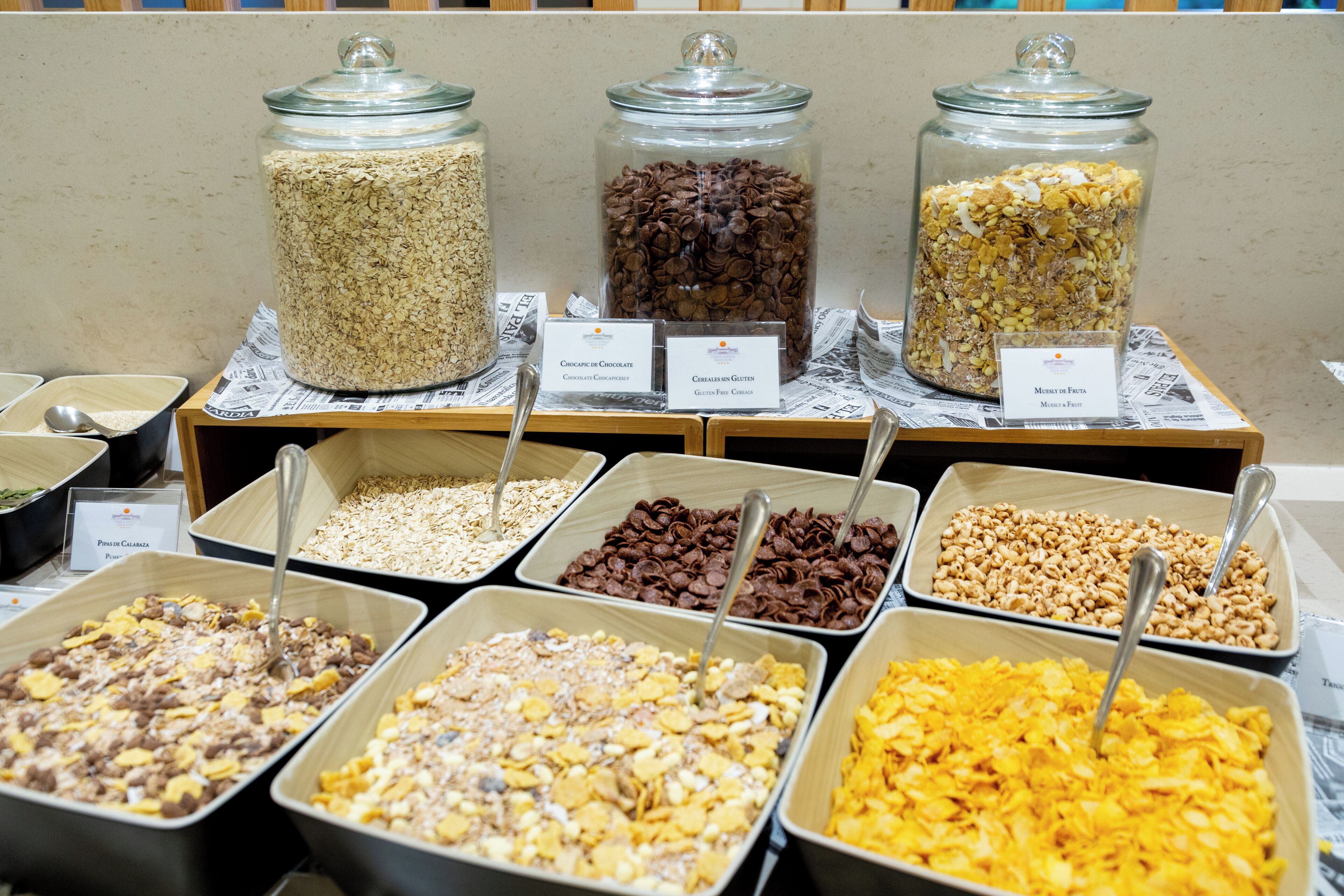 Un bufé de desayuno con diversas opciones de cereales y granola, presentados en frascos y bandejas, con sus ingredientes debidamente etiquetados.
