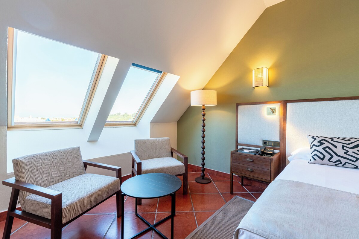 A hotel room with a bed, two armchairs, a small table, a nightstand, a lamp, and a view through skylights.