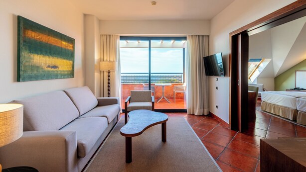 Een hotelkamer met een bank, een houten salontafel, een televisie, een balkon met uitzicht op zee en een schilderij aan de muur.