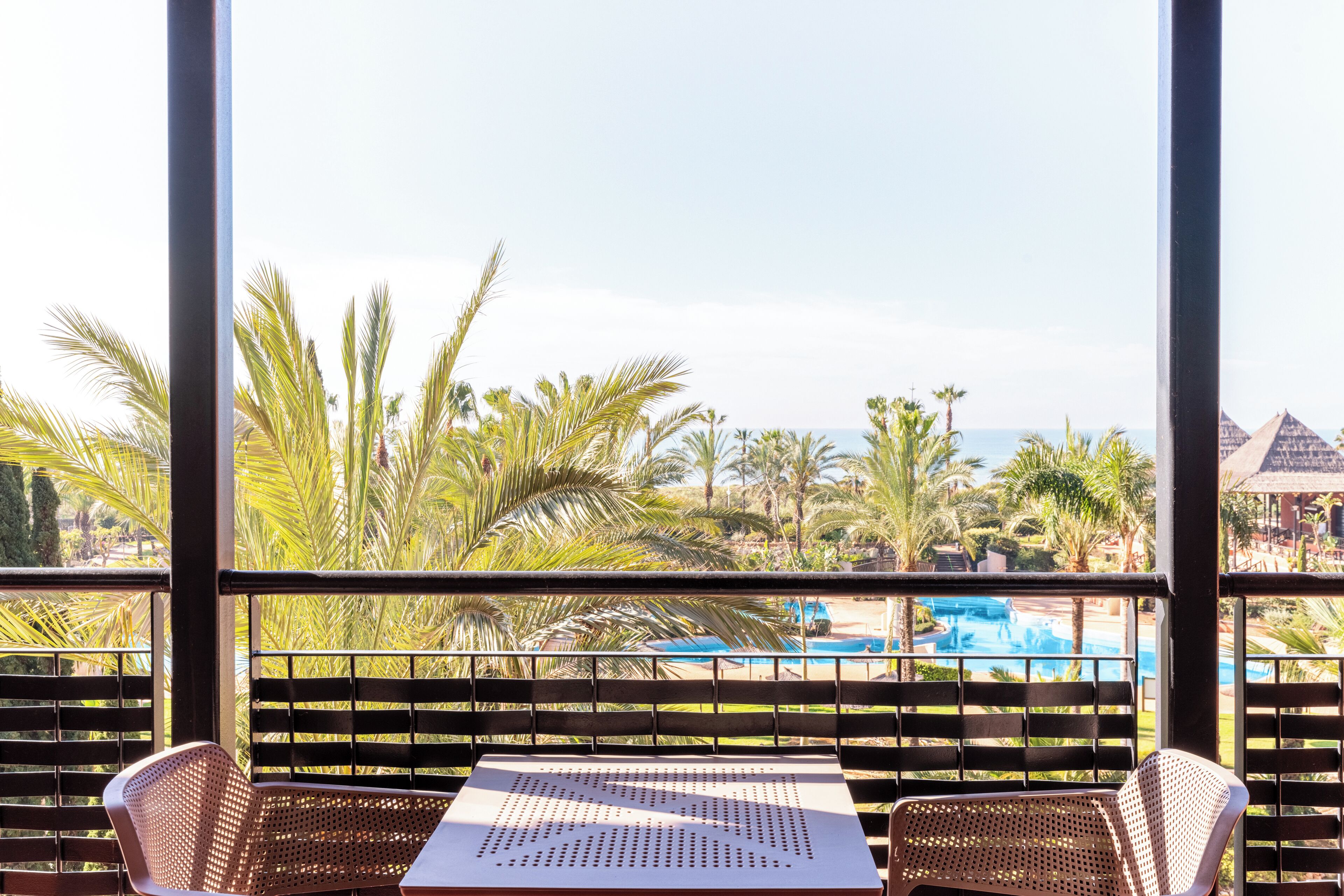 Un balcón con sillas de mimbre y una mesa, con vista a una piscina y palmeras.
