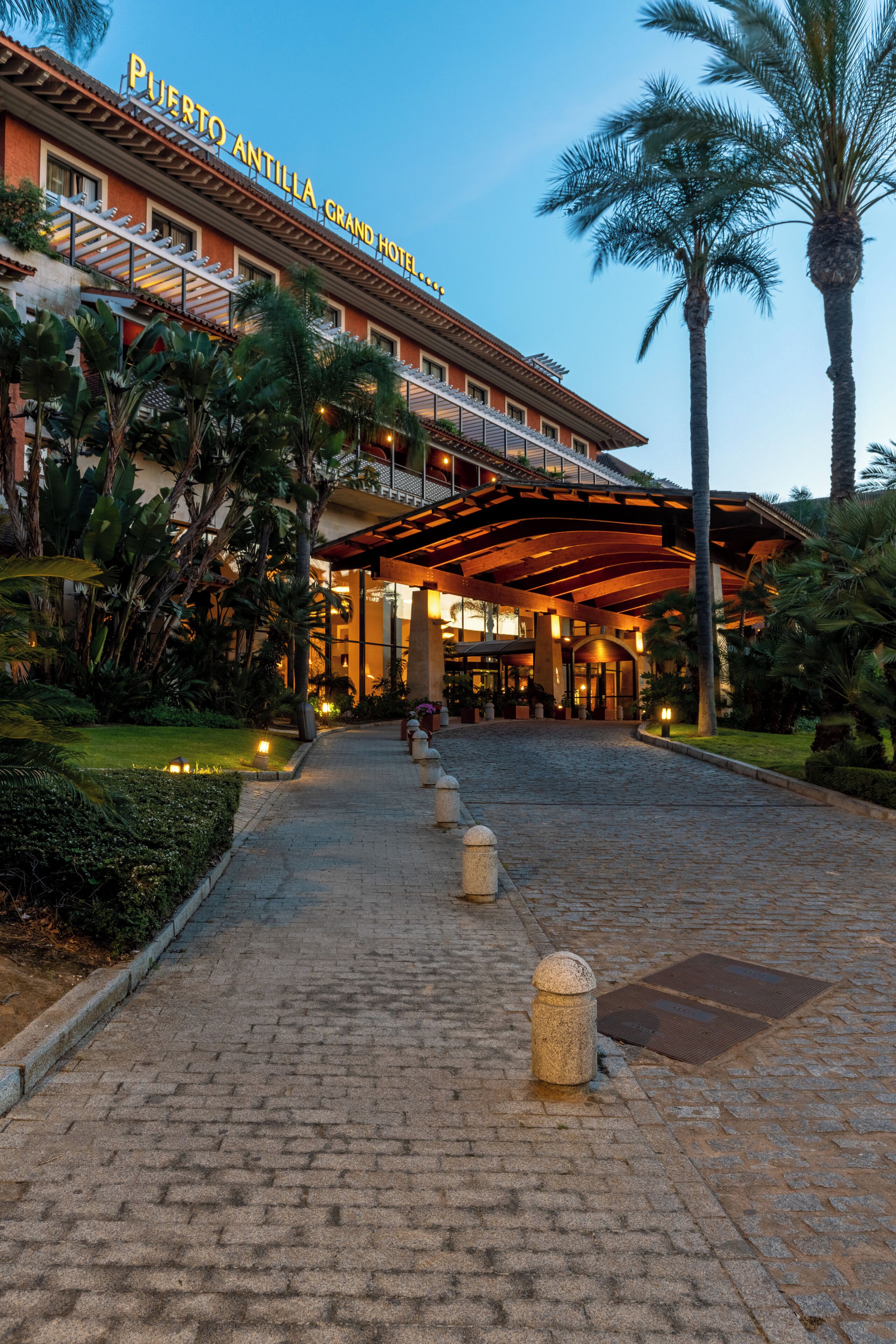 La entrada del Gran Hotel Puerto Antilla, con un sendero pavimentado, palmeras y un paseo techado.