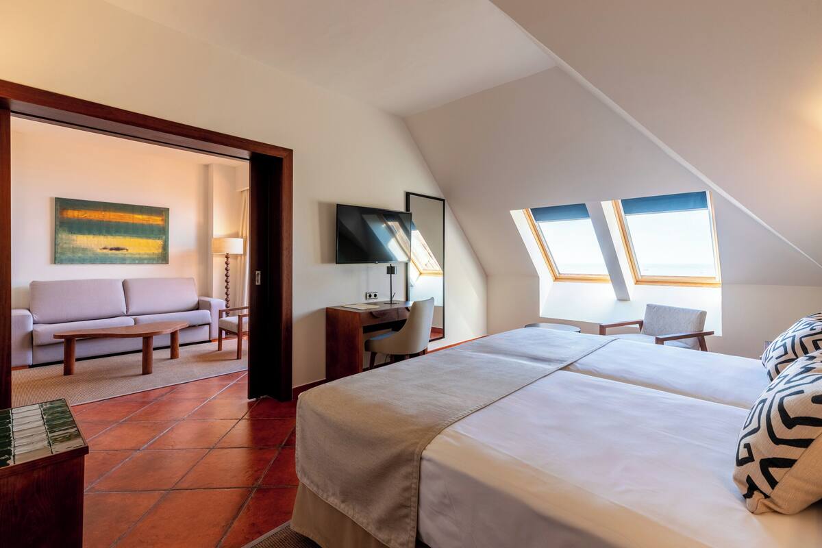 Een moderne hotelkamer met een groot bed, een bureau, een bank en een televisie. Er zijn dakramen en een schilderij aan de muur.