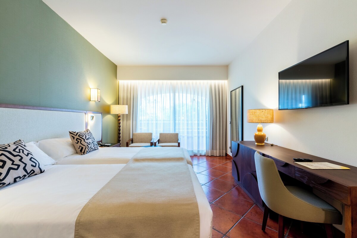 A hotel room with two beds, a desk, a chair, a TV, and a window with curtains.