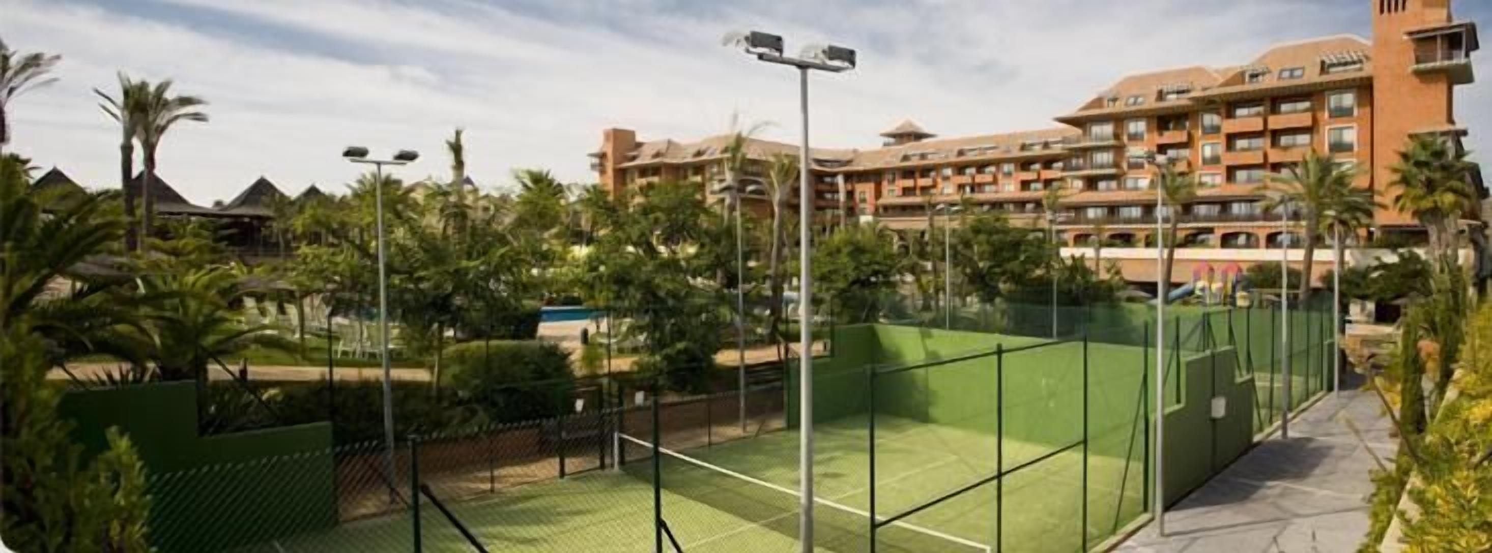 A tennis court surrounded by greenery and a hotel building with multiple balconies.