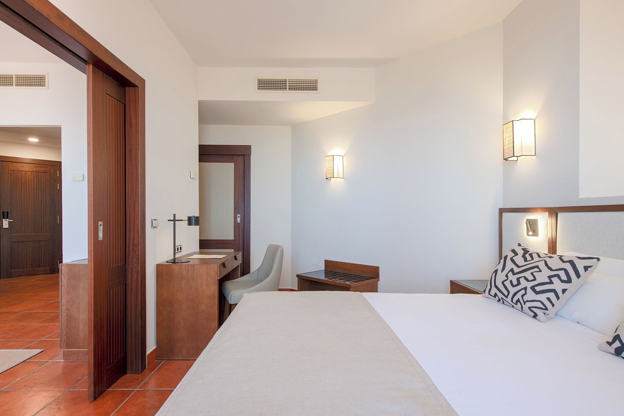 Quarto de hotel com cama, escrivaninha, cadeira, mesinha, espelho e banheiro com pia e vaso sanitário.