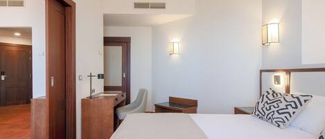 Quarto de hotel com cama, escrivaninha, cadeira, mesinha, espelho e banheiro com pia e vaso sanitário.