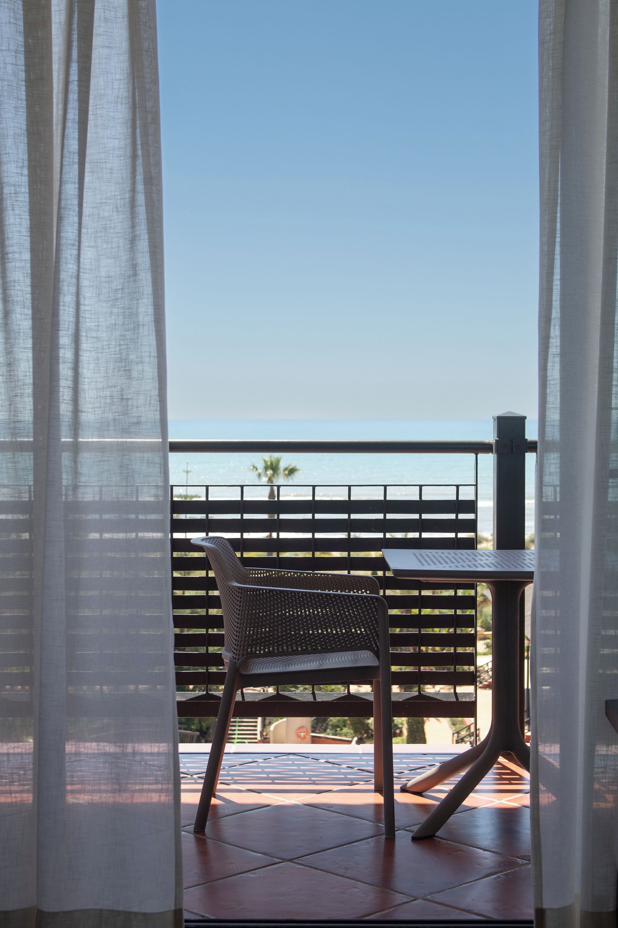 A balcony with a chair and table, overlooking a beach and palm trees.