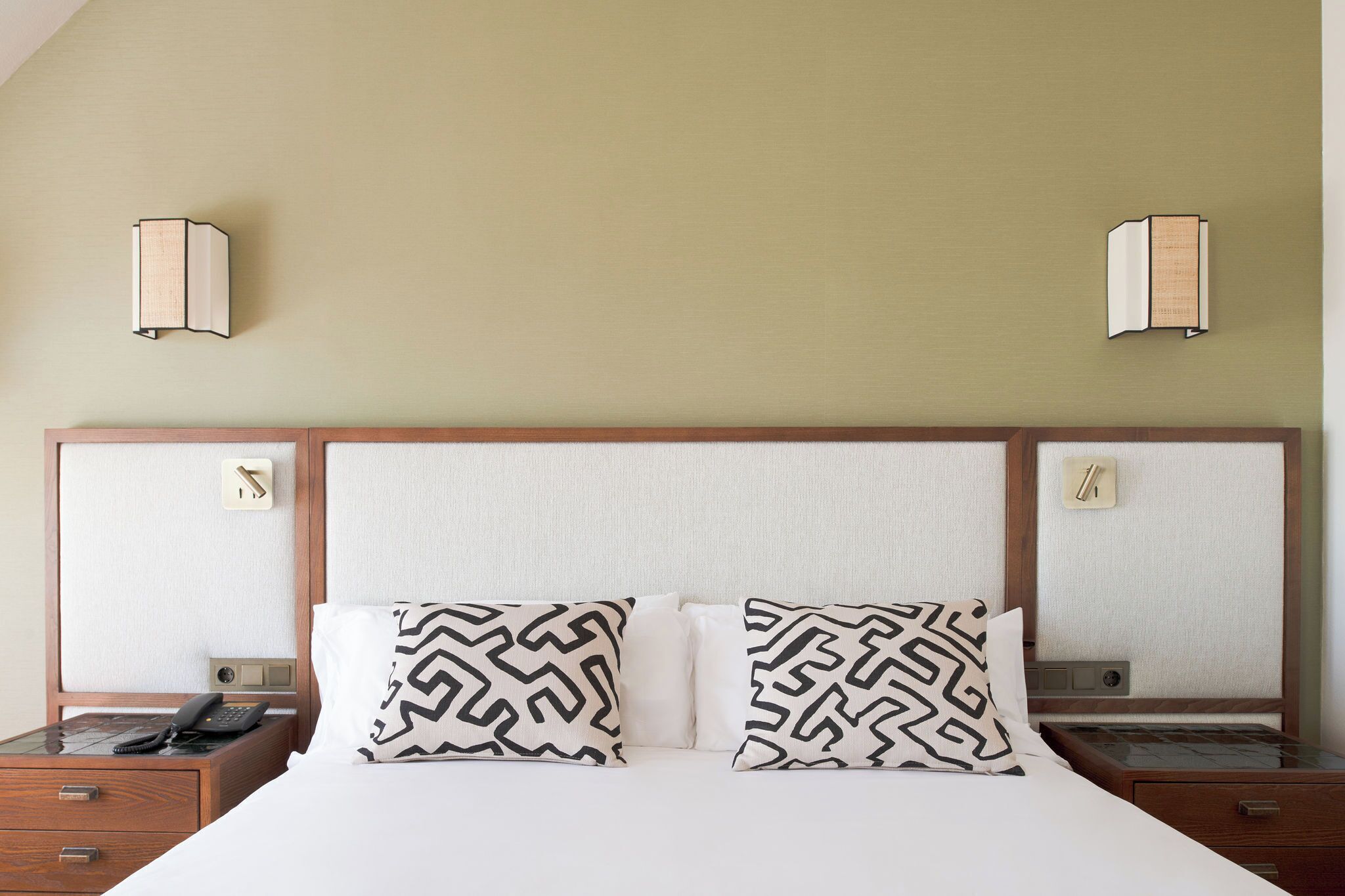 Een hotelkamer met een groot bed, twee gestreepte kussens, een nachtkastje met een telefoon en wandlampen.