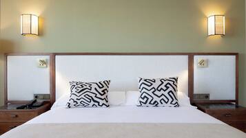 Quarto de hotel com uma cama grande, duas almofadas estampadas, mesas de cabeceira com abajures e uma cabeceira de madeira.