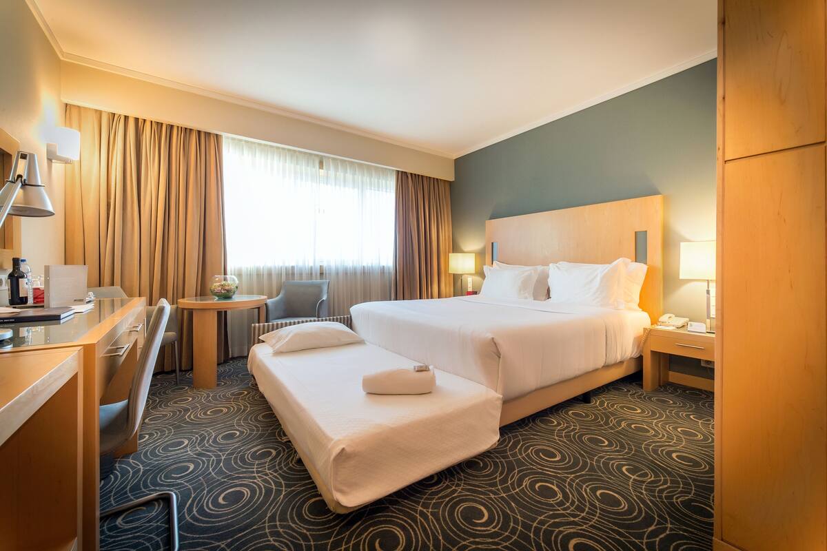 Ein Hotelzimmer mit Bett, Schreibtisch, Stuhl, einem kleinen Tisch und einem Nachttisch mit Lampe.