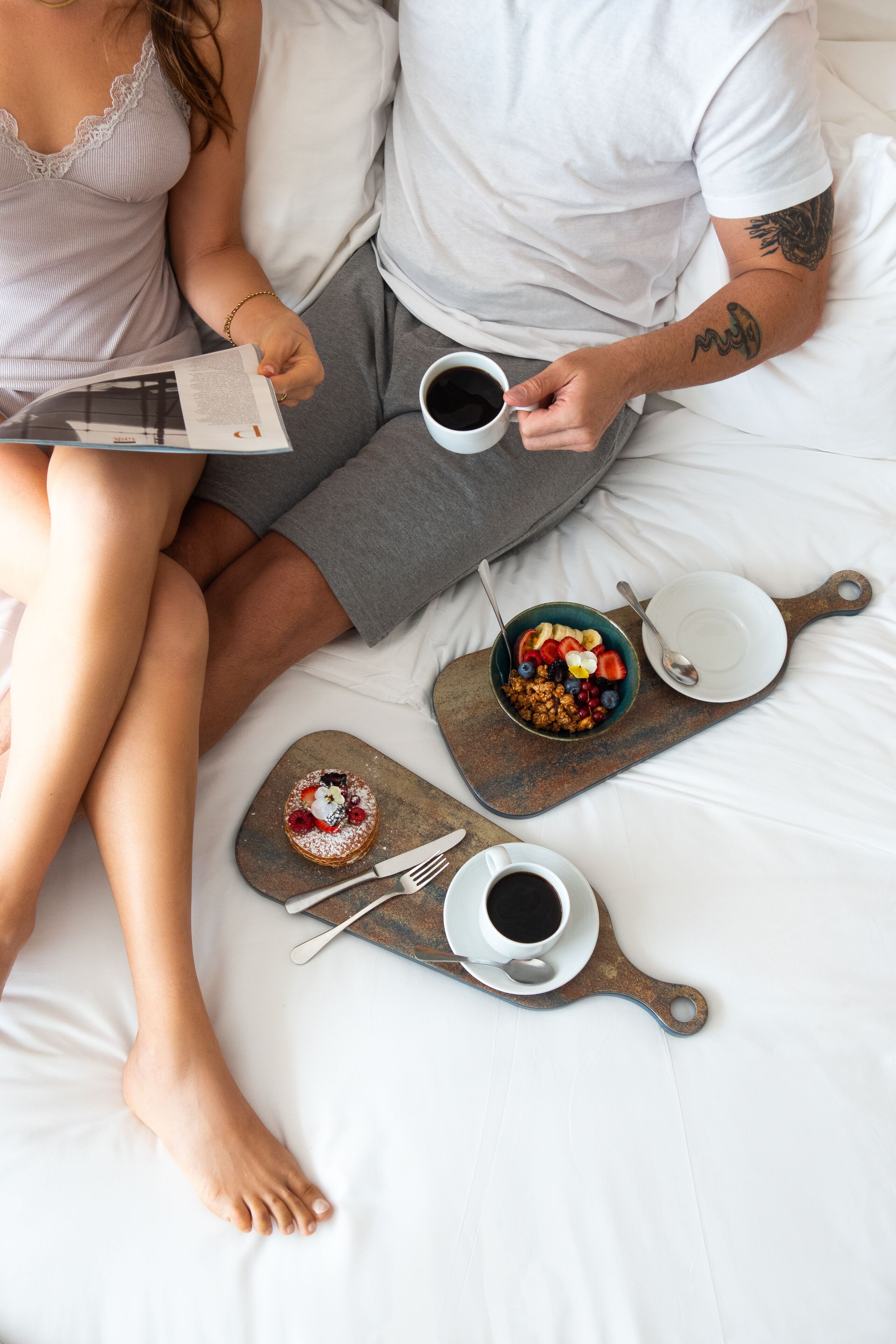 Een koppel in een bed met ontbijtartikelen, waaronder een kom cornflakes, een gebakje en twee kopjes koffie.